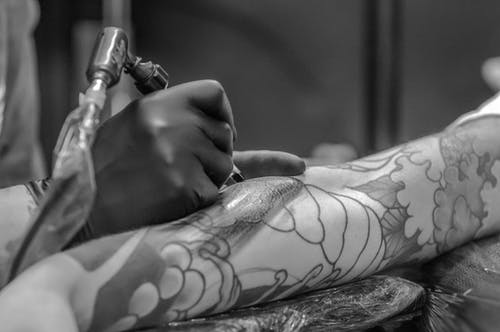 На фотографии изображен процесс создания татуировки. Рука некоего человека видна со стороны, а на ней видна процесс нанесения татуировки. Искусственные иглы и краски используются для тщательного нанесения изображения на кожу. Татуировка, еще не завершенная, начинает появляться на руке, создавая уникальный искусственный узор.