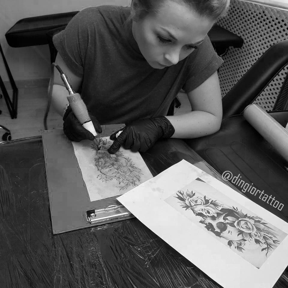 На снимке изображена женщина, использующая тату-машинку для того, чтобы татуировать искусственную кожу. В ее руках находится машинка, которая внимательно следит за контурами и деталями рисунка. Женщина уделяет особое внимание перерисовке картинки с бумаги на искусственной коже, придавая изображению уникальность и художественный характер.